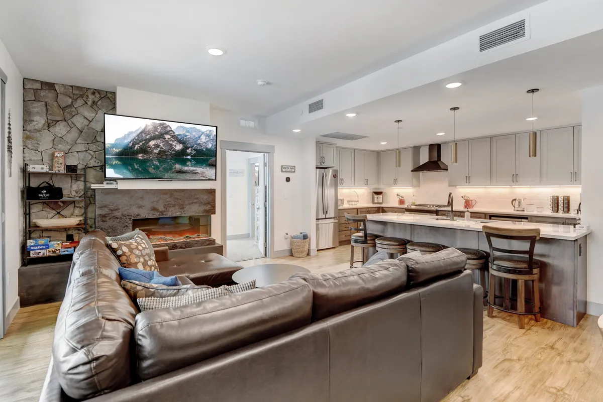 This image presents a spacious open-plan living area that seamlessly integrates a leather sectional sofa, a large wall-mounted TV, a striking stone fireplace, and the kitchen island with bar stools in the background. The arrangement showcases comfortable seating for group gatherings, the inviting warmth of the fireplace, and easy access to both dining and kitchen spaces.