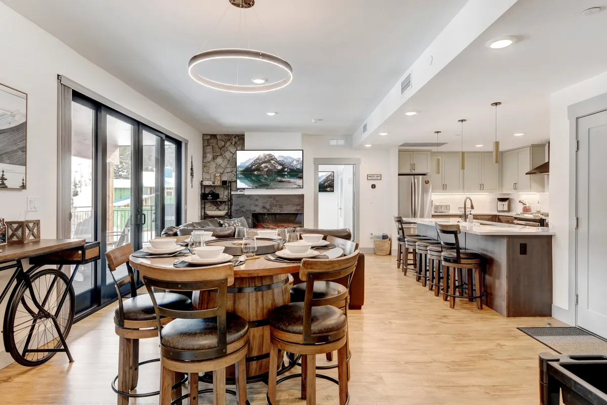 A welcoming dining and kitchen space is featured, with a large dining table set for communal meals, a kitchen island for casual gatherings, and a stone fireplace providing ambience. Large windows offer glimpses of the mountain views outside, enhancing the sense of warmth and togetherness.
