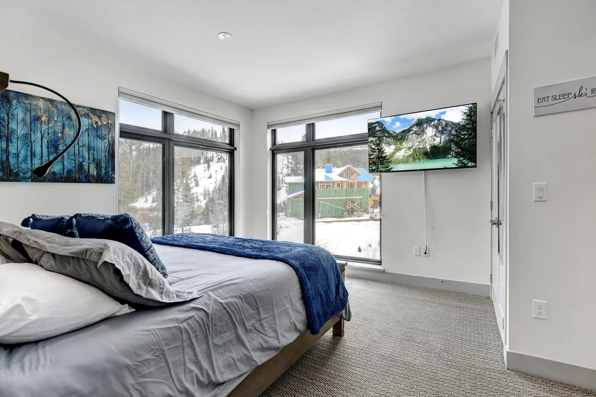 A tranquil master bedroom featuring a plush bed with blue throw blanket, large windows offering mountain views, a wall-mounted TV, and calming artwork, creating an inviting space for rest and relaxation.