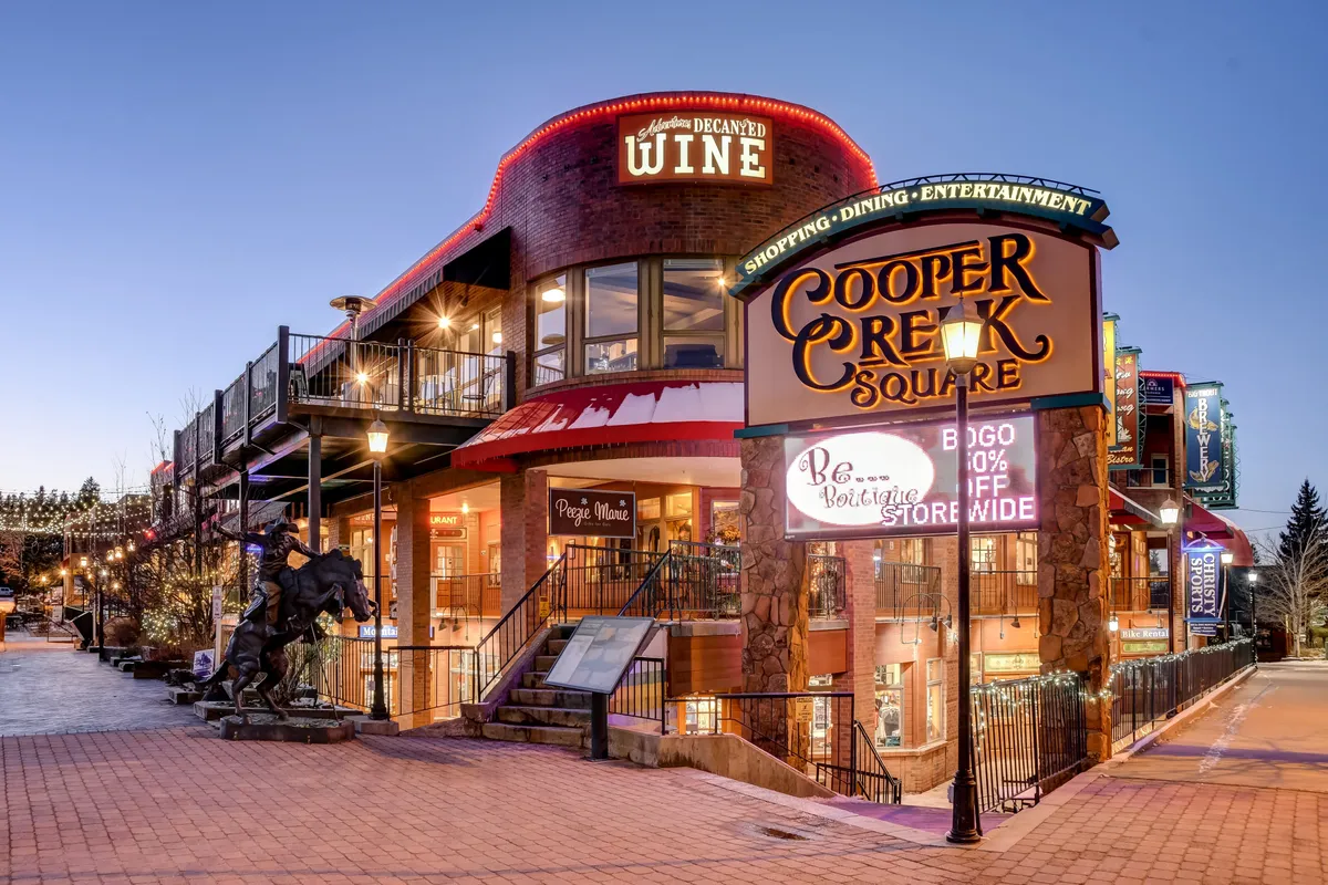A lively scene at Cooper Creek Square in downtown Winter Park, featuring a prominent sign, decorative wine signage, an outdoor dining area bustling with patrons, and a statue of a horse. The space is designed for both relaxation and socializing, with a clear atmosphere of activity and community.