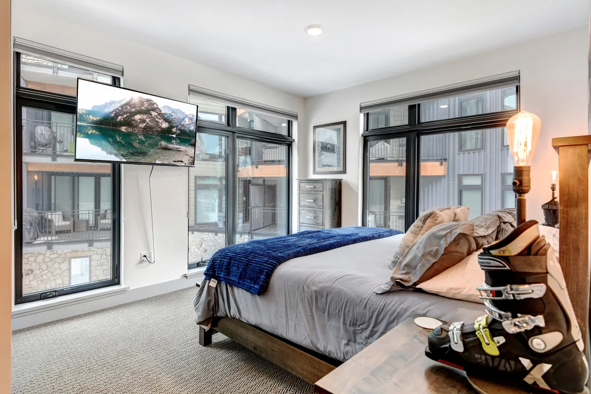 An inviting bedroom scene with a plush bed draped in a blue throw blanket, ski boots resting on a bedside table, and a mounted TV displaying a mountain vista. The room exudes both coziness and a sense of adventure, ideal for relaxing after a day on the slopes.