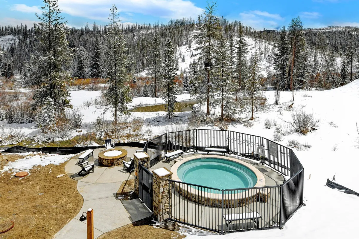An inviting outdoor scene showcasing a steaming hot tub and a glowing fire pit surrounded by snow, set within a wooded landscape. The area is designed for both relaxation and social gatherings, with enough space for groups to enjoy the amenities together under the open sky.