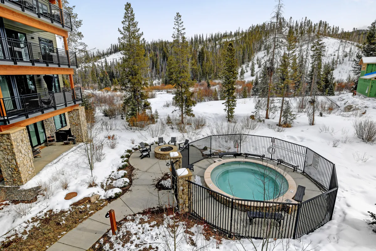 A shared outdoor amenity area featuring a bubbling hot tub and a large fire pit surrounded by chairs, set amid a snowy forest landscape. The area exudes relaxation and invites guests to soak after a day of adventure or gather for stories by the fire under the stars.
