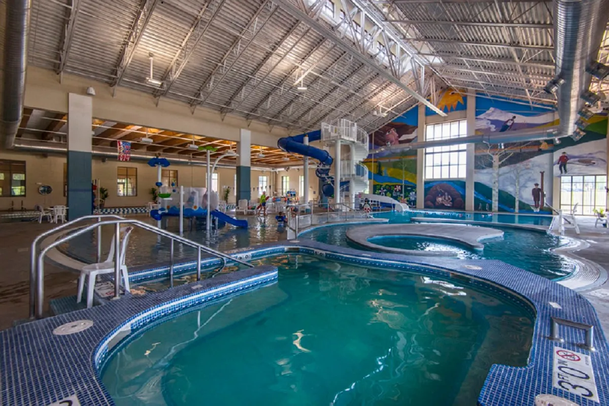 A lively indoor recreation center scene featuring a large swimming pool, a colorful waterslide, artistic wall murals, and ample lounge seating, all bathed in natural light. Children and families can be imagined enjoying water play and relaxation together.