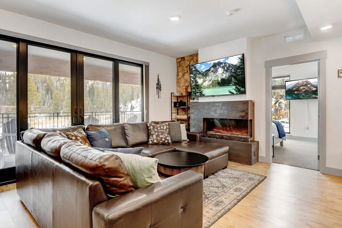 The image showcases a spacious, stylish living room with a plush leather sectional sofa arranged around a glowing fireplace. Large windows frame stunning views of snow-capped mountains, while TVs display additional scenic vistas. The setting is cozy and perfect for relaxing after outdoor adventures, inviting guests to unwind with family and friends.
