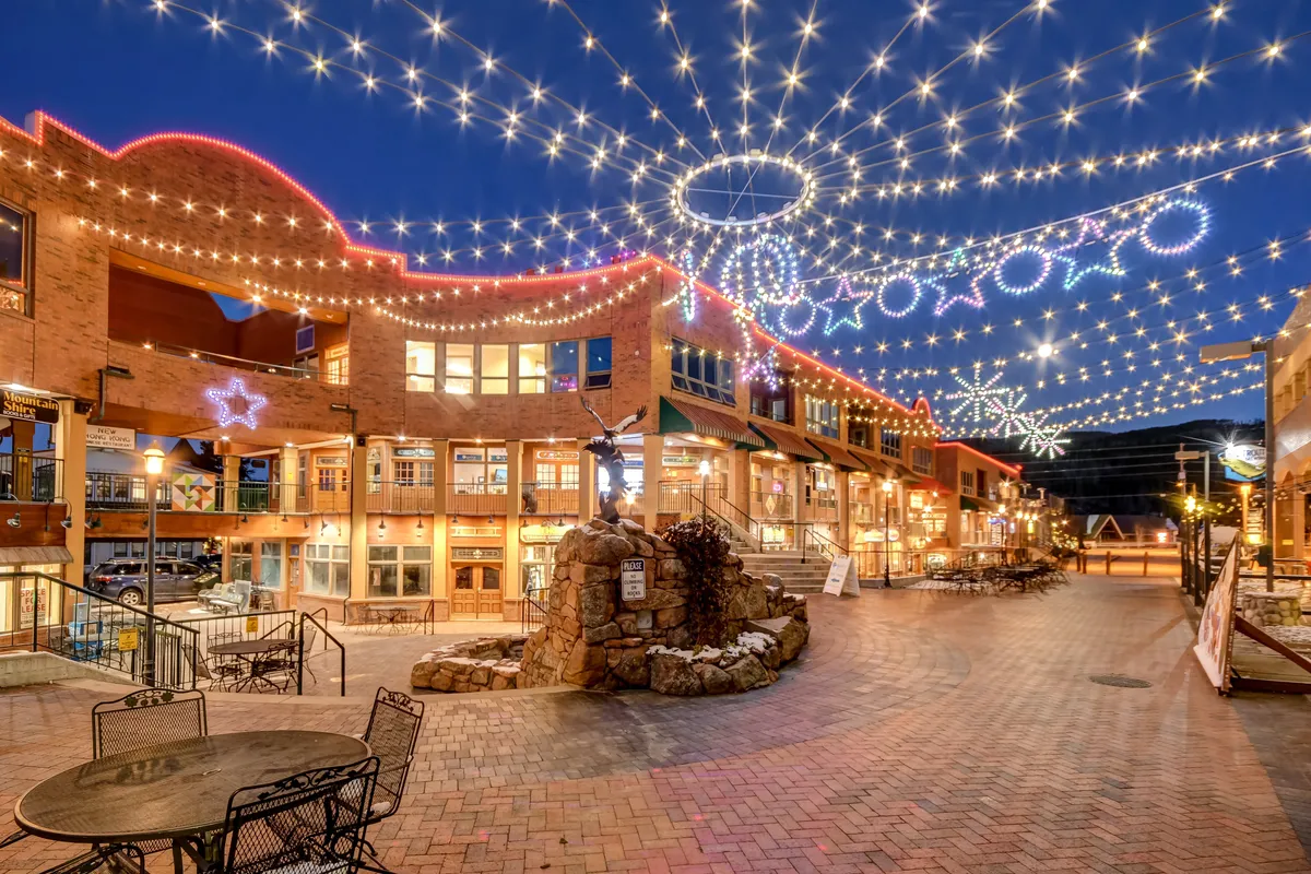 A festive nighttime streetscape featuring decorative string lights, a central sculpture, and building facades set up for dining and shopping. The atmosphere is joyful and communal, suggesting an inviting place for evening outings and social events.